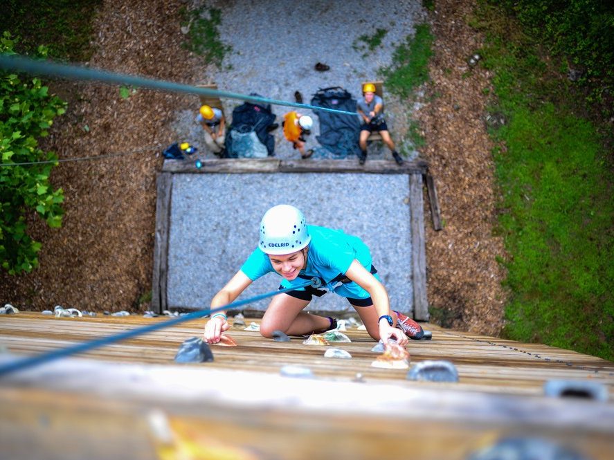A smiling camper in a blue helmet climbing the wall, viewed from above