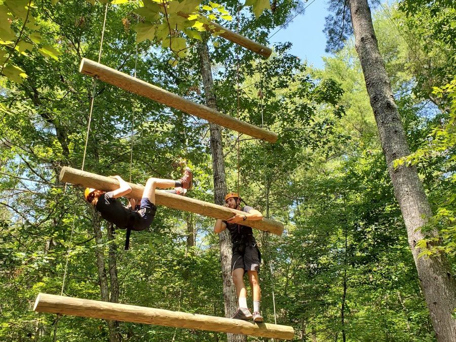 A camper in a helmet and harness navigating a ropes course in the trees