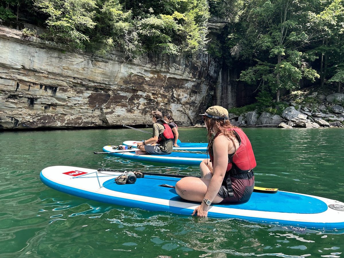 Campers in life jackets on paddleboards beside a rocky riverbank