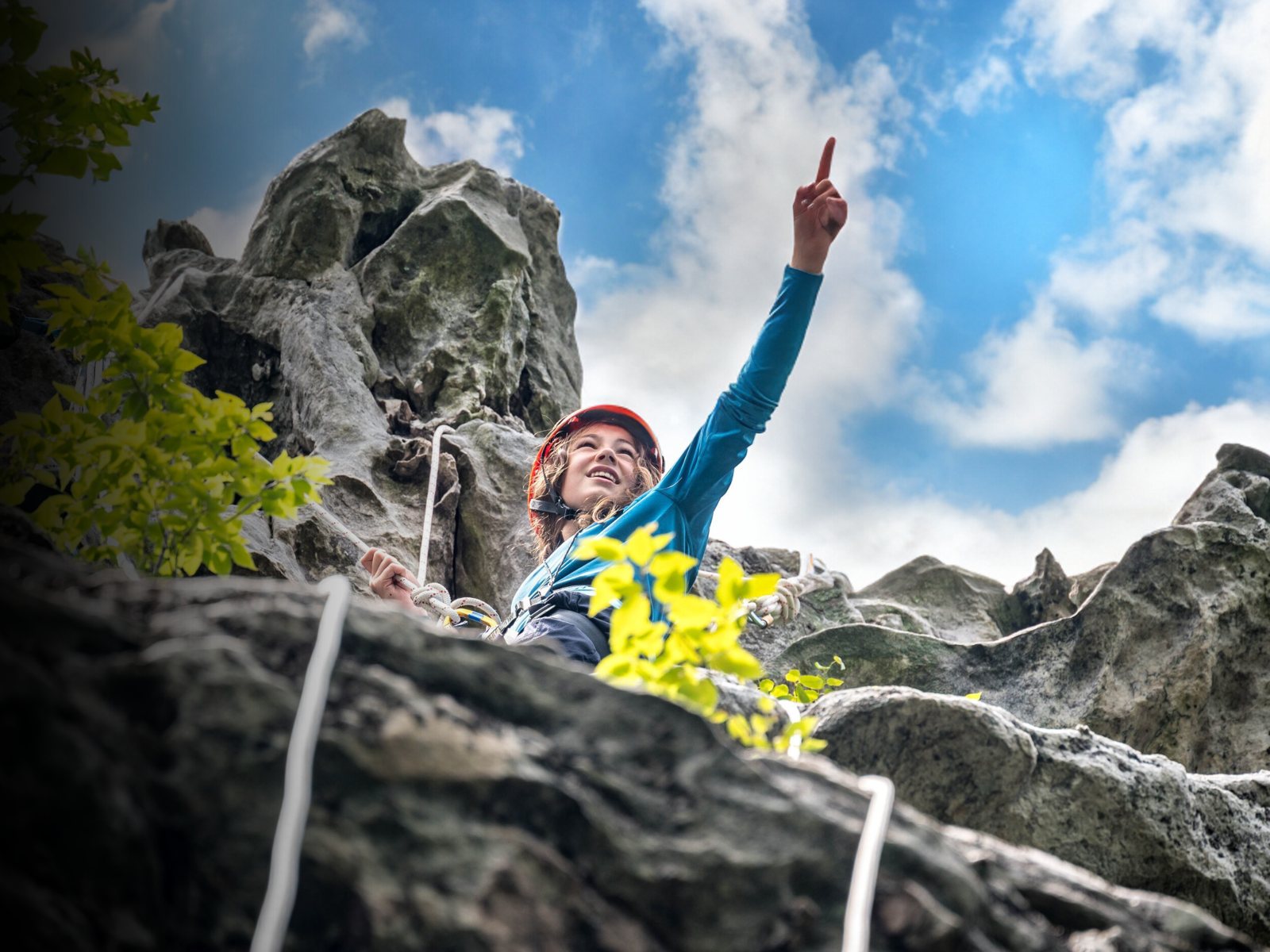 A helmeted climber pointing up a rock face beneath a blue sky