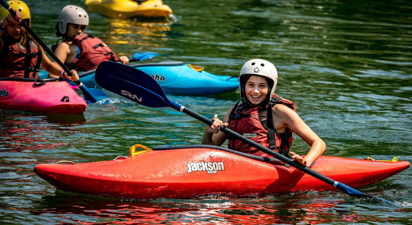 Helmeted campers paddling brightly colored kayaks on the water