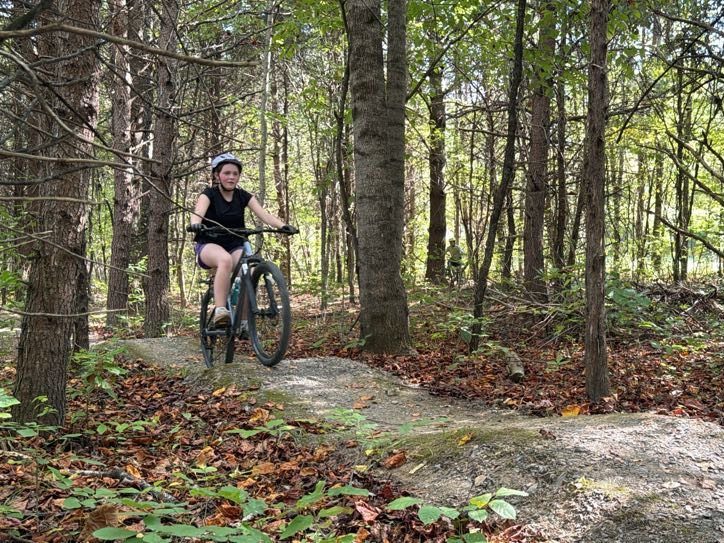 A camper riding a mountain bike over a rock feature on a wooded trail at Base Camp