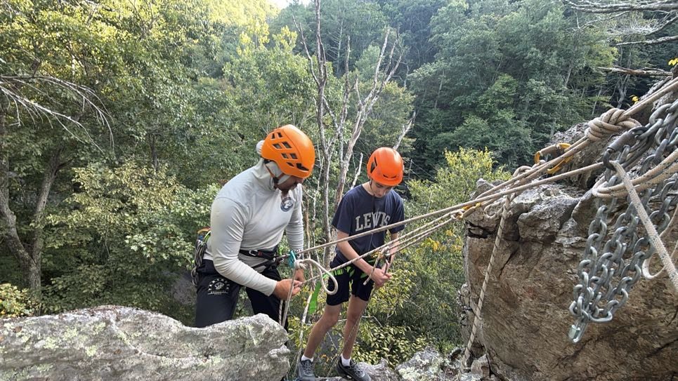 An instructor and a helmeted student checking ropes beside a rock ledge