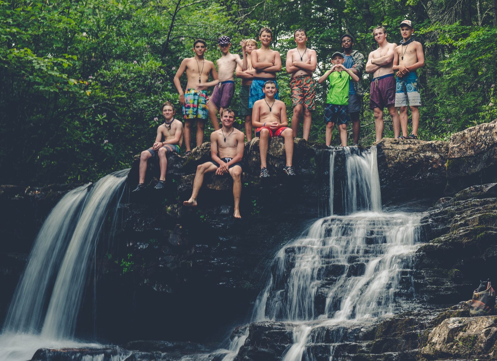 Campers standing and sitting on a rock ledge at the top of a waterfall in the woods
