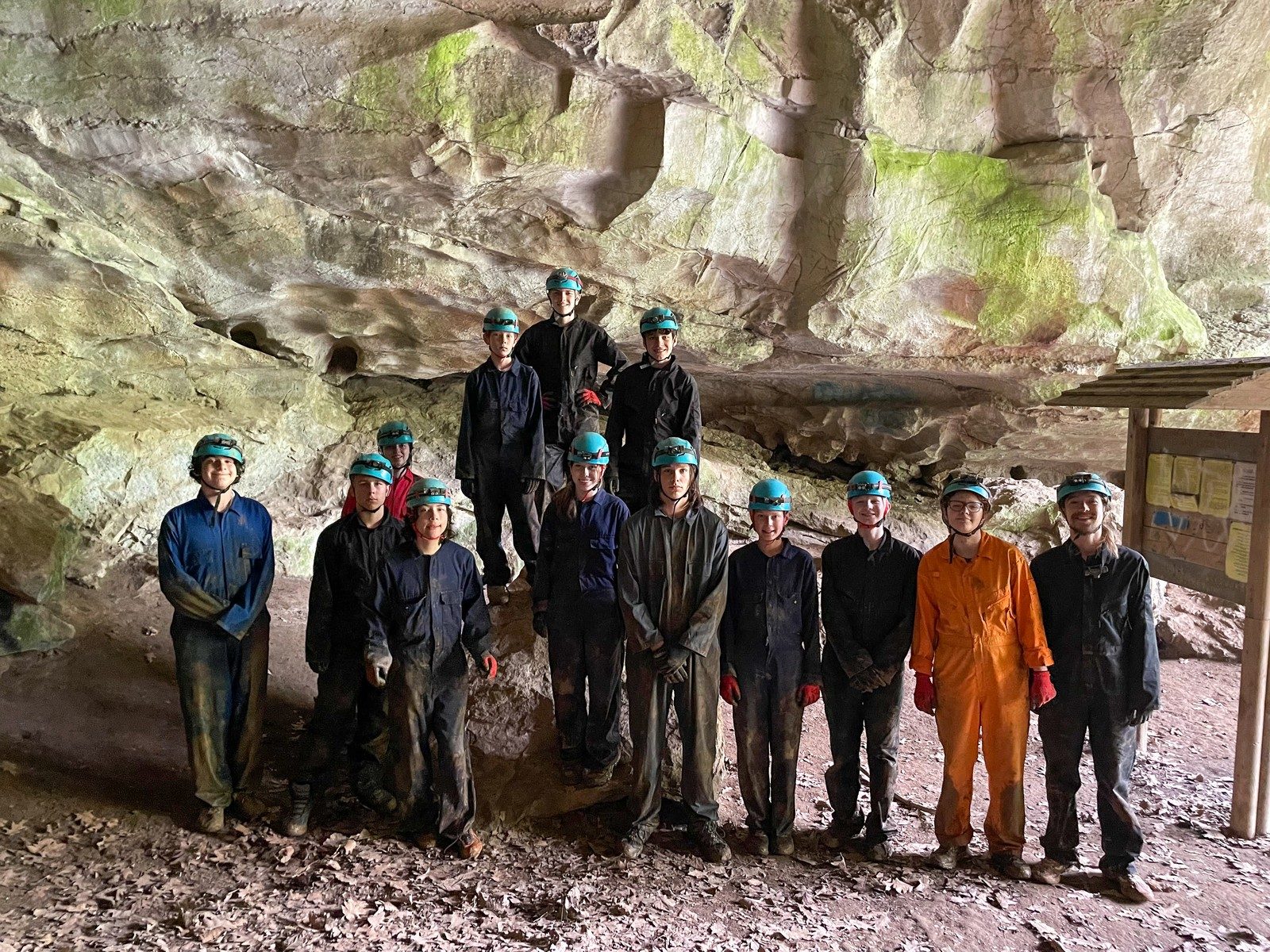 A caving group in coveralls and helmets lined up under a rock overhang
