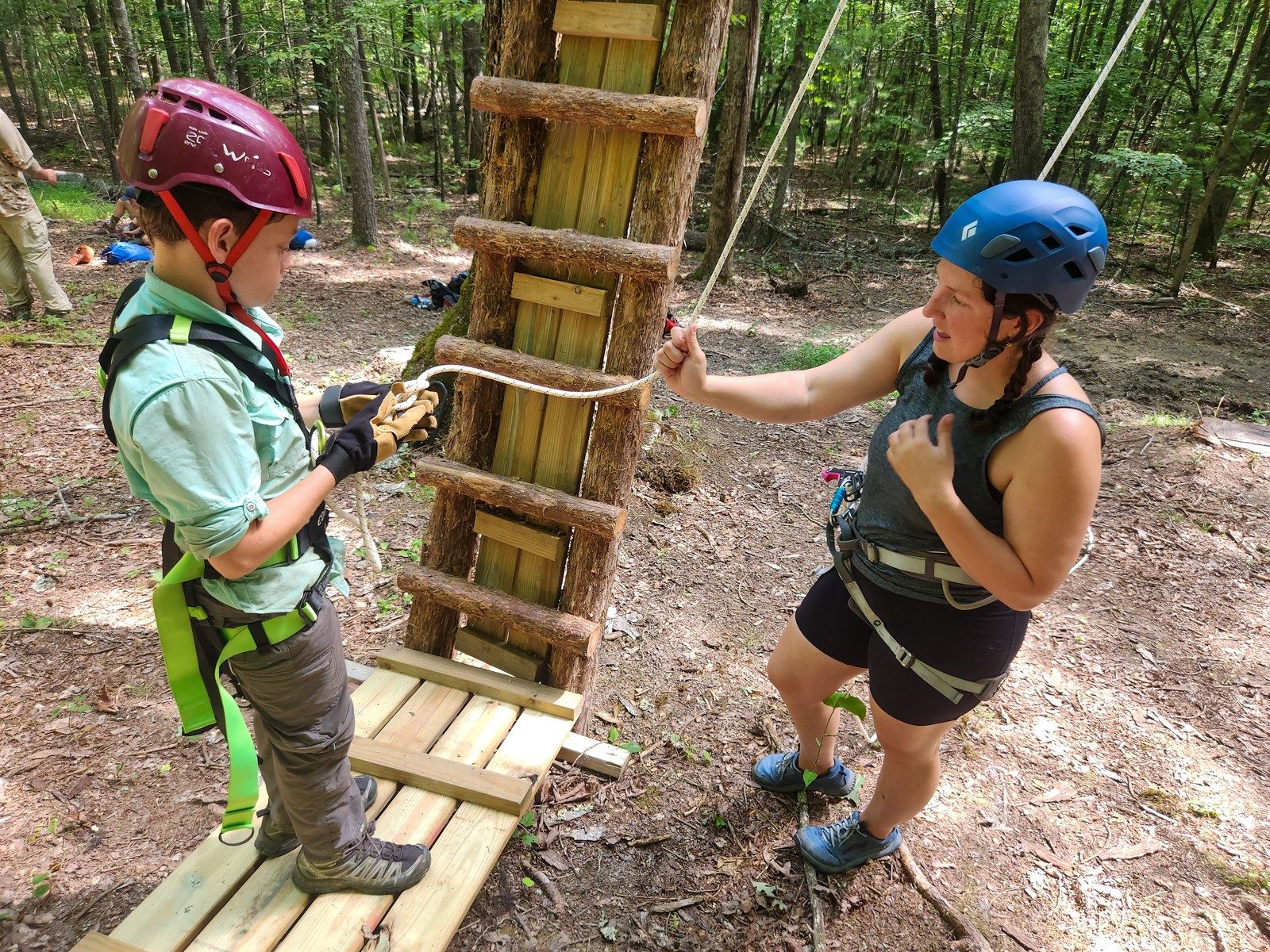 A counselor checking a young camper's harness at the foot of the ropes course ladder